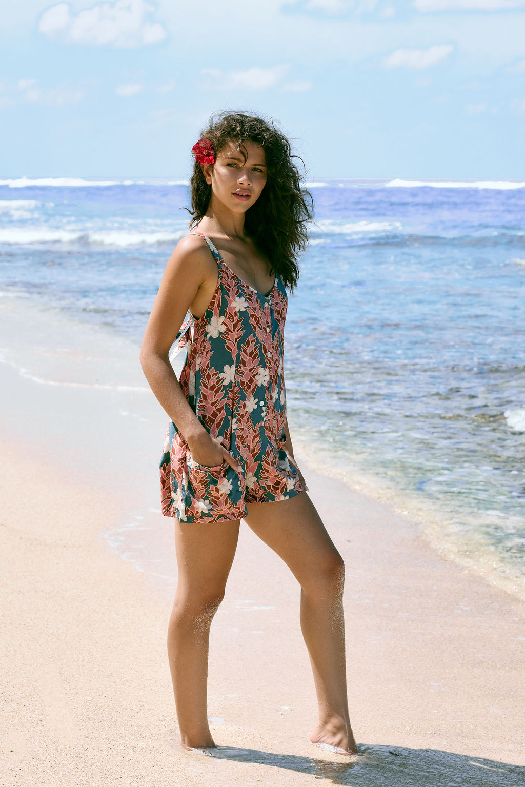 woman with brown hair wearing a blue thin strap tie back romper with red ginger and tiare print. 