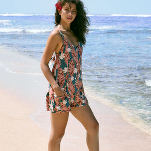 woman with brown hair wearing a blue thin strap tie back romper with red ginger and tiare print. 