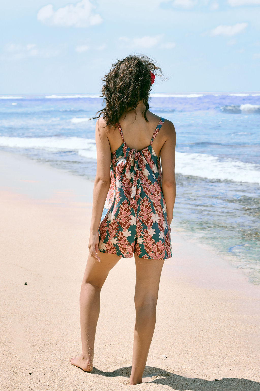 woman with brown hair wearing a blue thin strap tie back romper with red ginger and tiare print. 