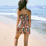 woman with brown hair wearing a blue thin strap tie back romper with red ginger and tiare print. 