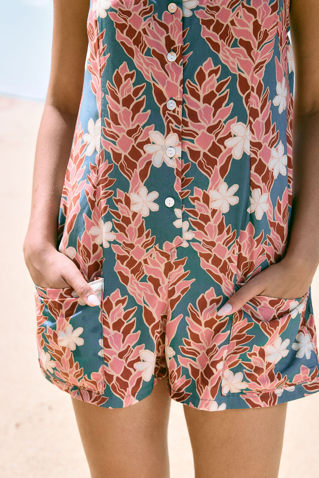 woman with brown hair wearing a blue thin strap tie back romper with red ginger and tiare print. 