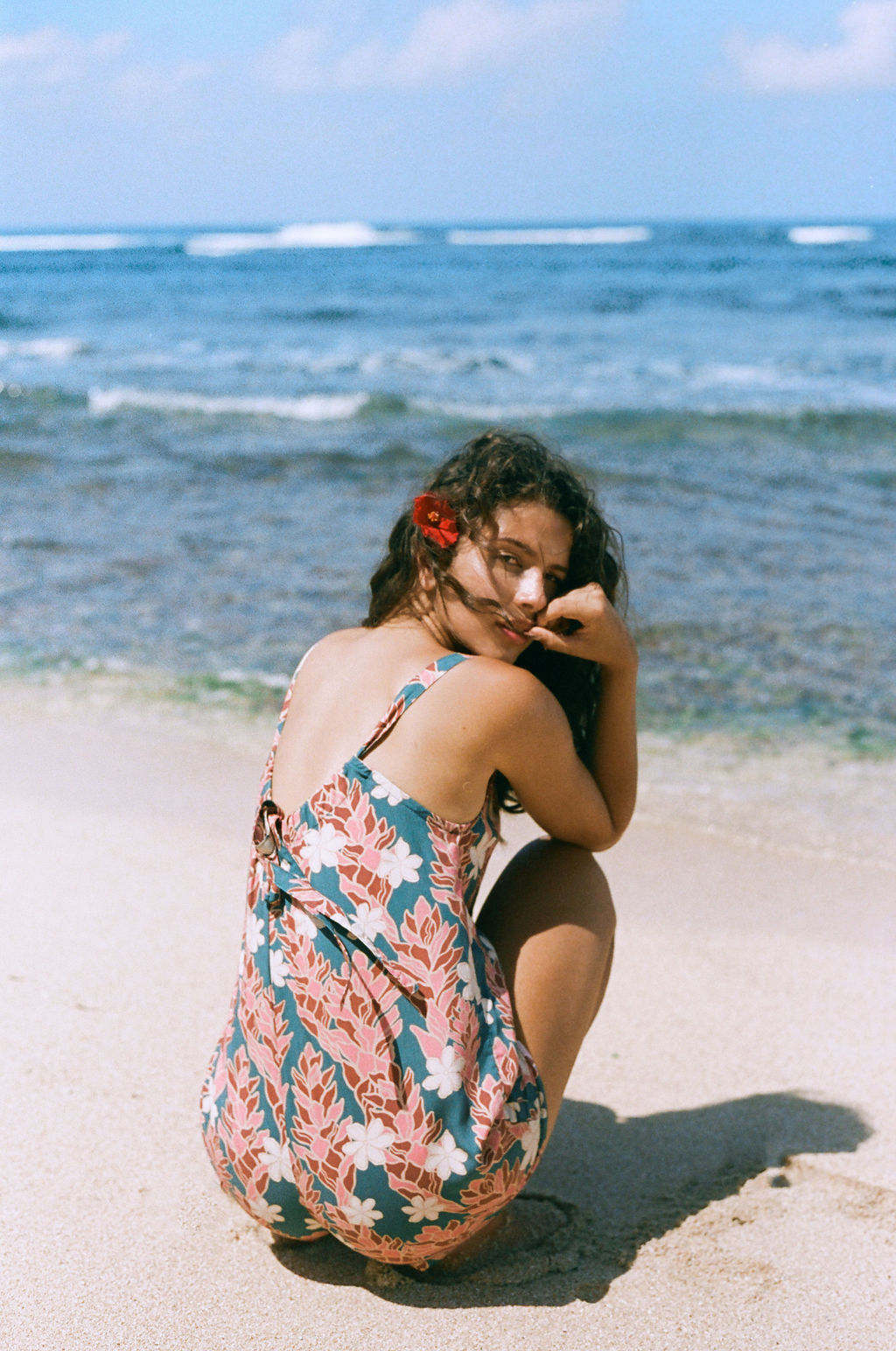woman with brown hair wearing a blue thin strap tie back romper with red ginger and tiare print. 