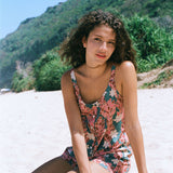 woman with brown hair wearing a blue thin strap tie back romper with red ginger and tiare print. 