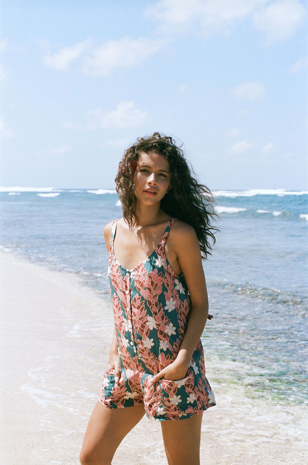 woman with brown hair wearing a blue thin strap tie back romper with red ginger and tiare print. 