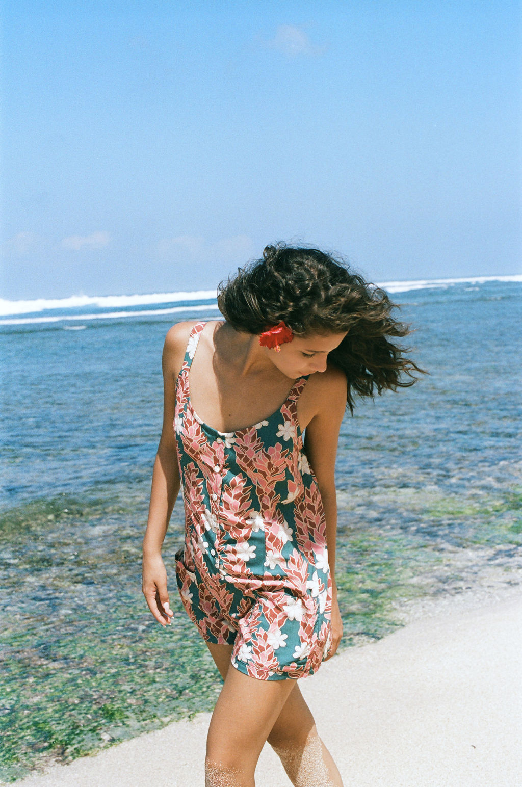 woman with brown hair wearing a blue thin strap tie back romper with red ginger and tiare print. 