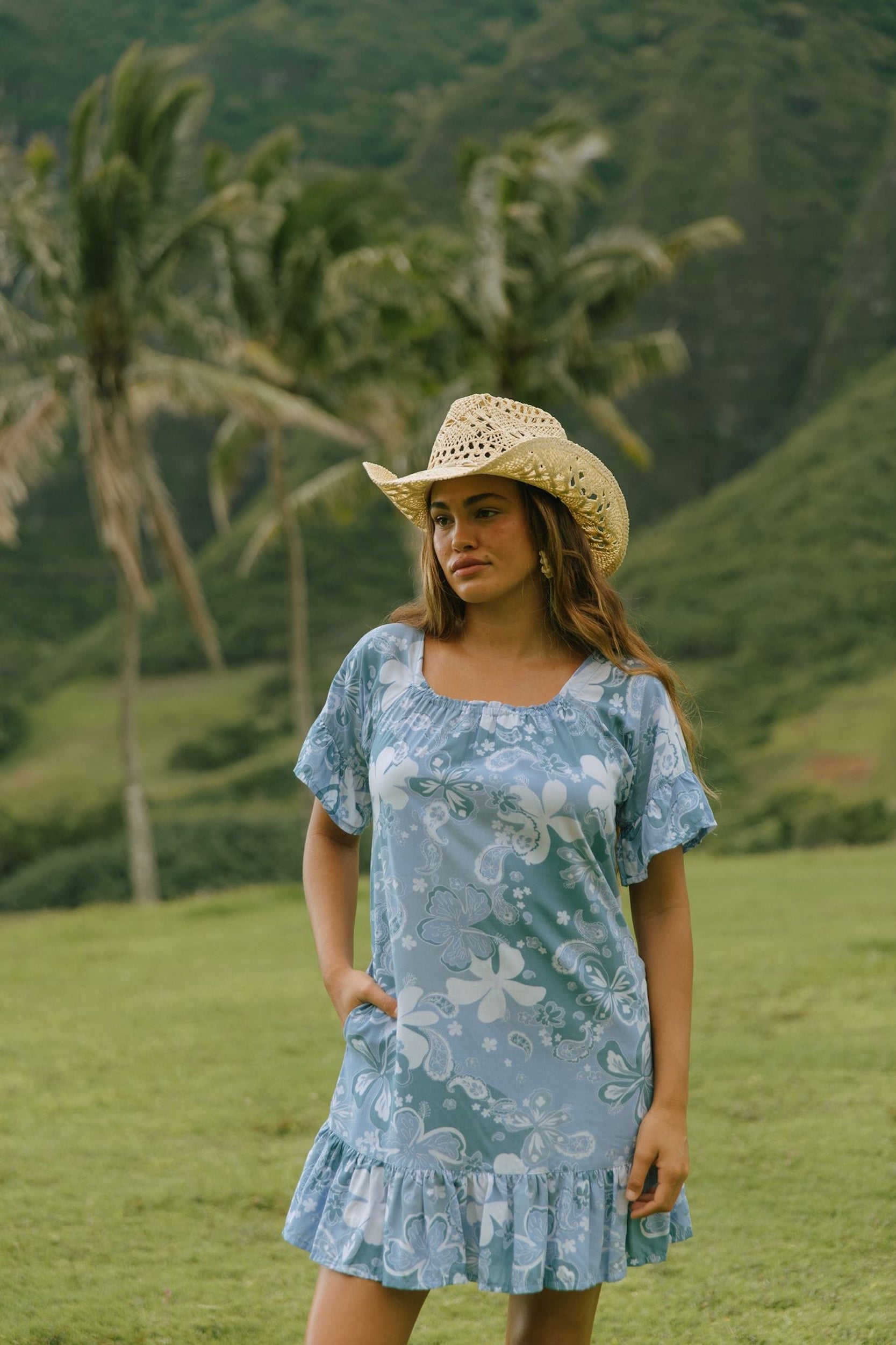 woman with brown hair wearing a blue paniolo printed mu'u style dress with flutter sleeves and a ruffle hem 