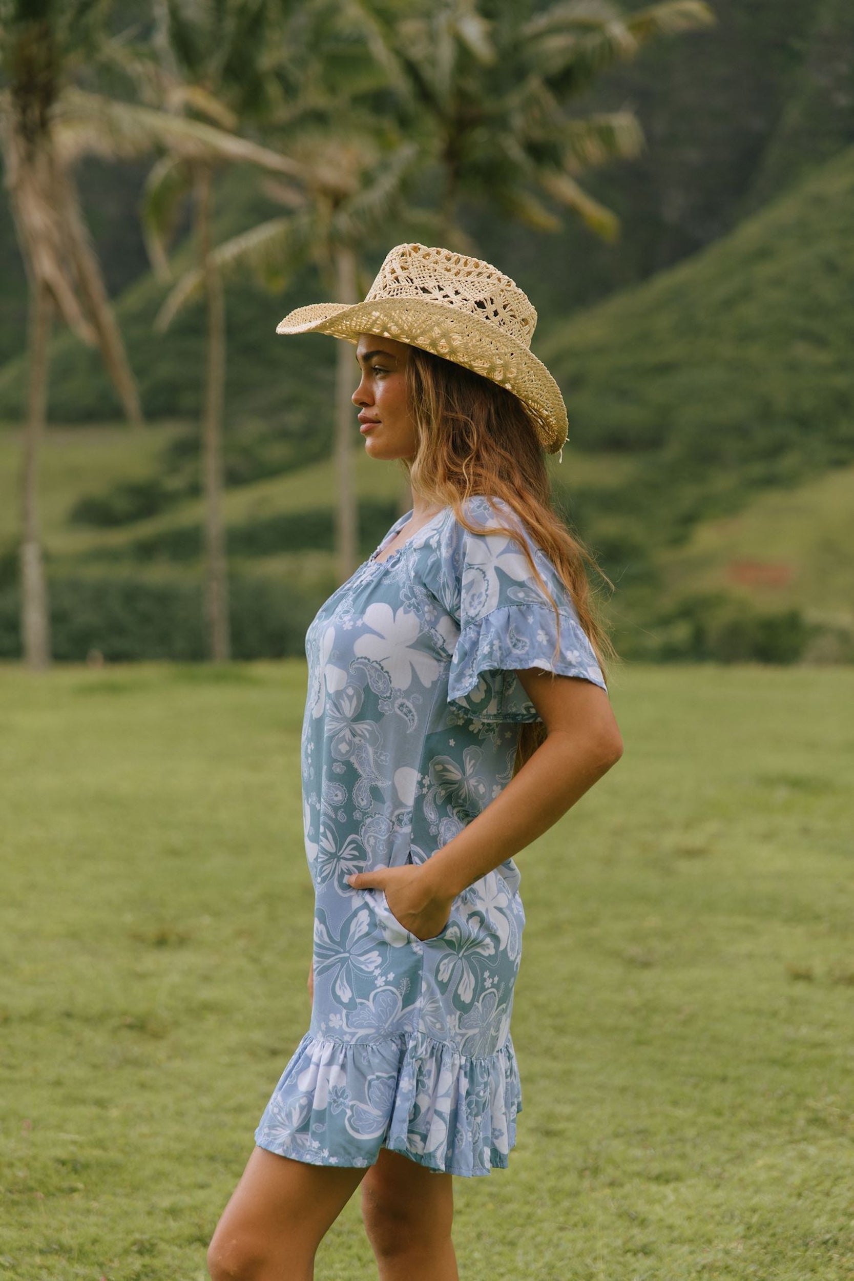 woman with brown hair wearing a blue paniolo printed mu'u style dress with flutter sleeves and a ruffle hem 