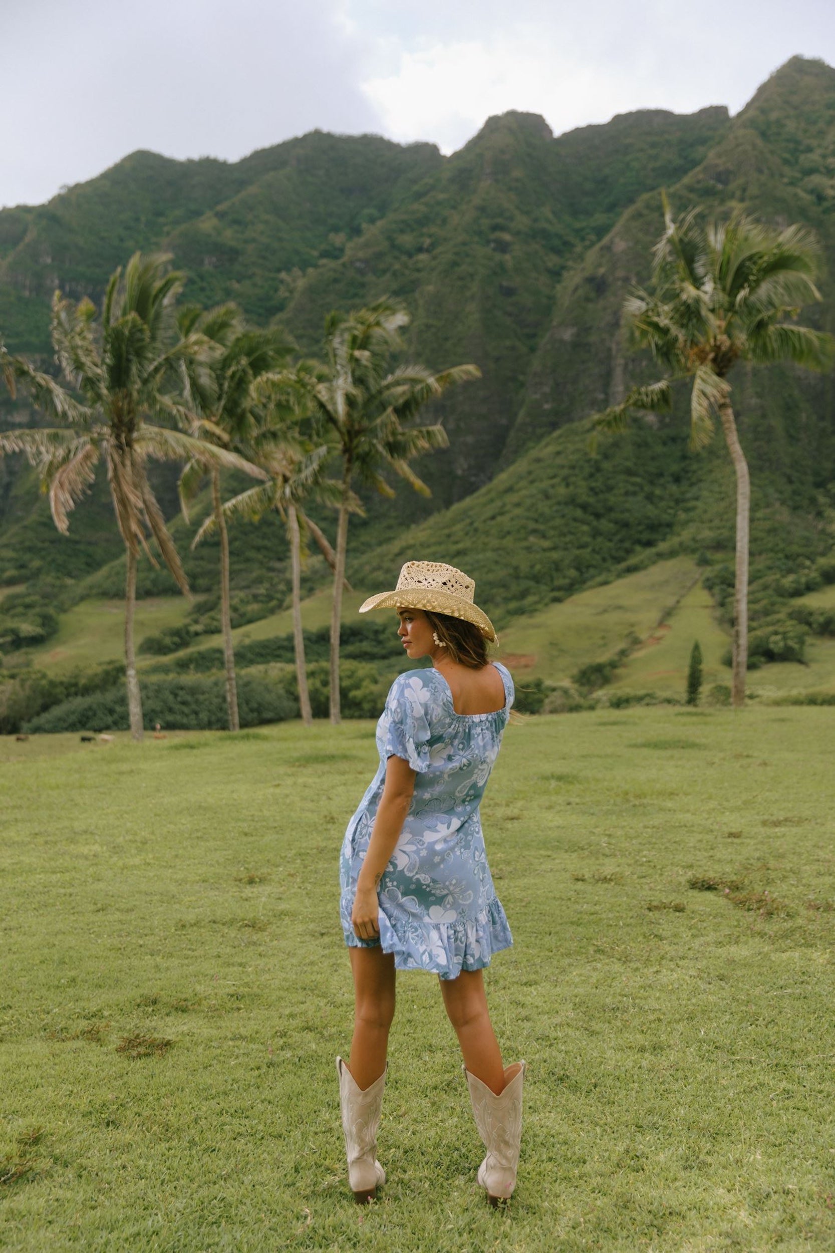 woman with brown hair wearing a blue paniolo printed mu'u style dress with flutter sleeves and a ruffle hem 