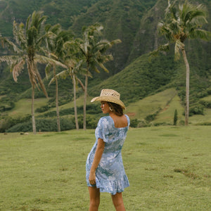woman with brown hair wearing a blue paniolo printed mu'u style dress with flutter sleeves and a ruffle hem 