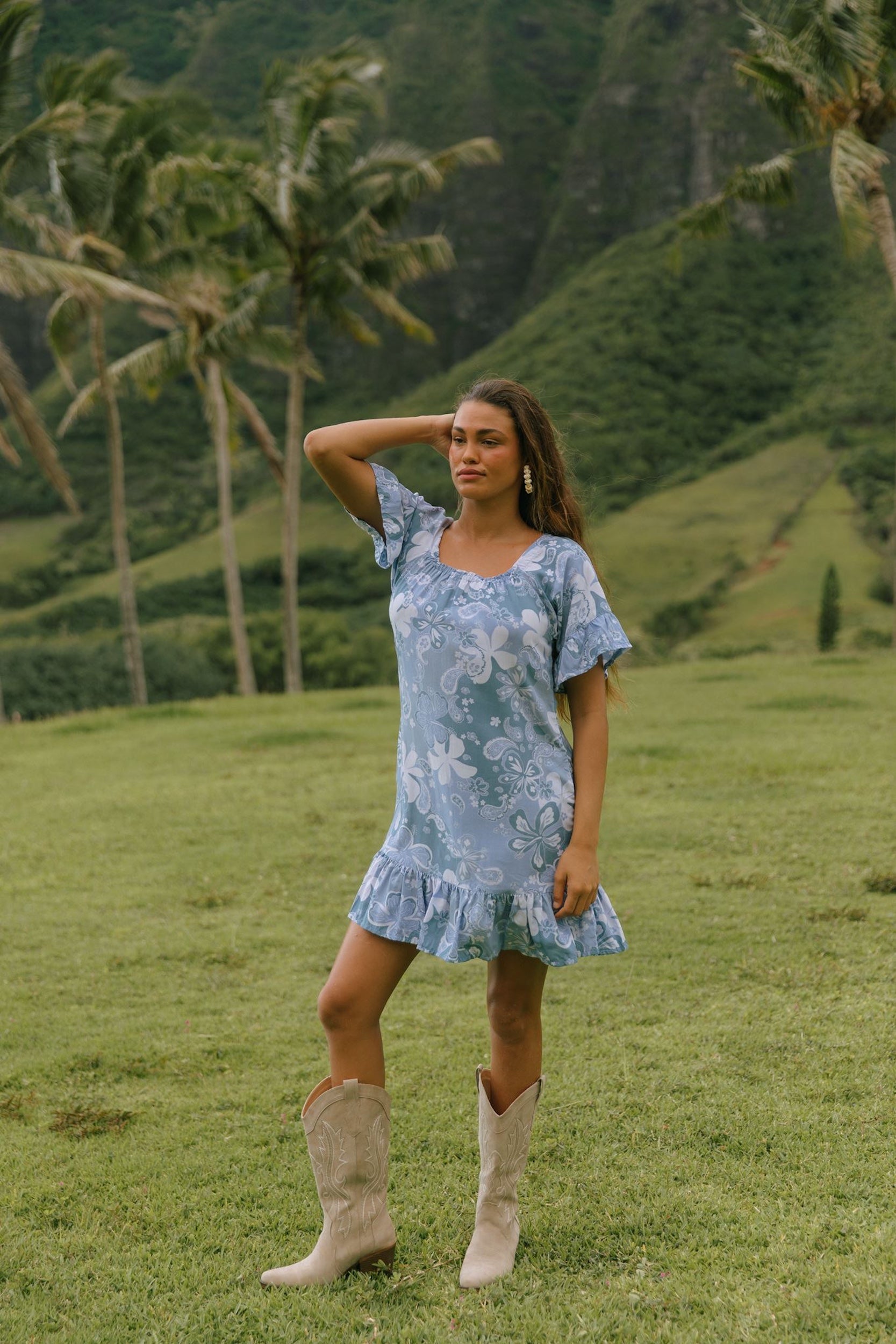 woman with brown hair wearing a blue paniolo printed mu'u style dress with flutter sleeves and a ruffle hem 