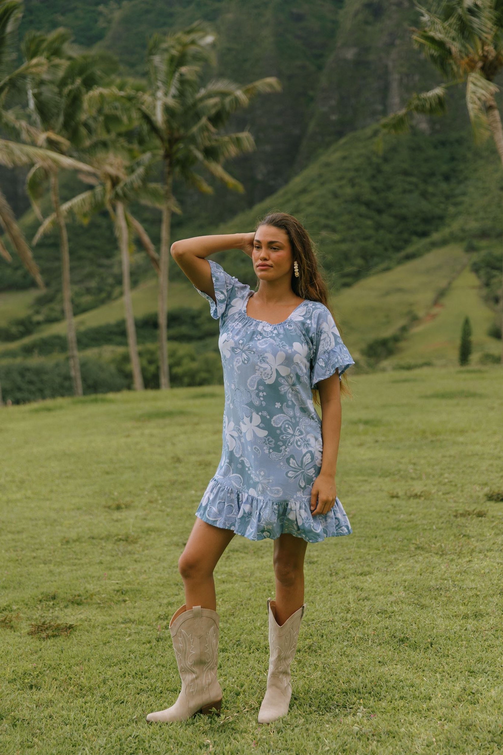 woman with brown hair wearing a blue paniolo printed mu'u style dress with flutter sleeves and a ruffle hem 