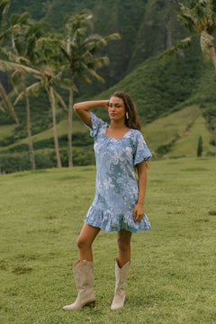 woman with brown hair wearing a blue paniolo printed mu'u style dress with flutter sleeves and a ruffle hem 