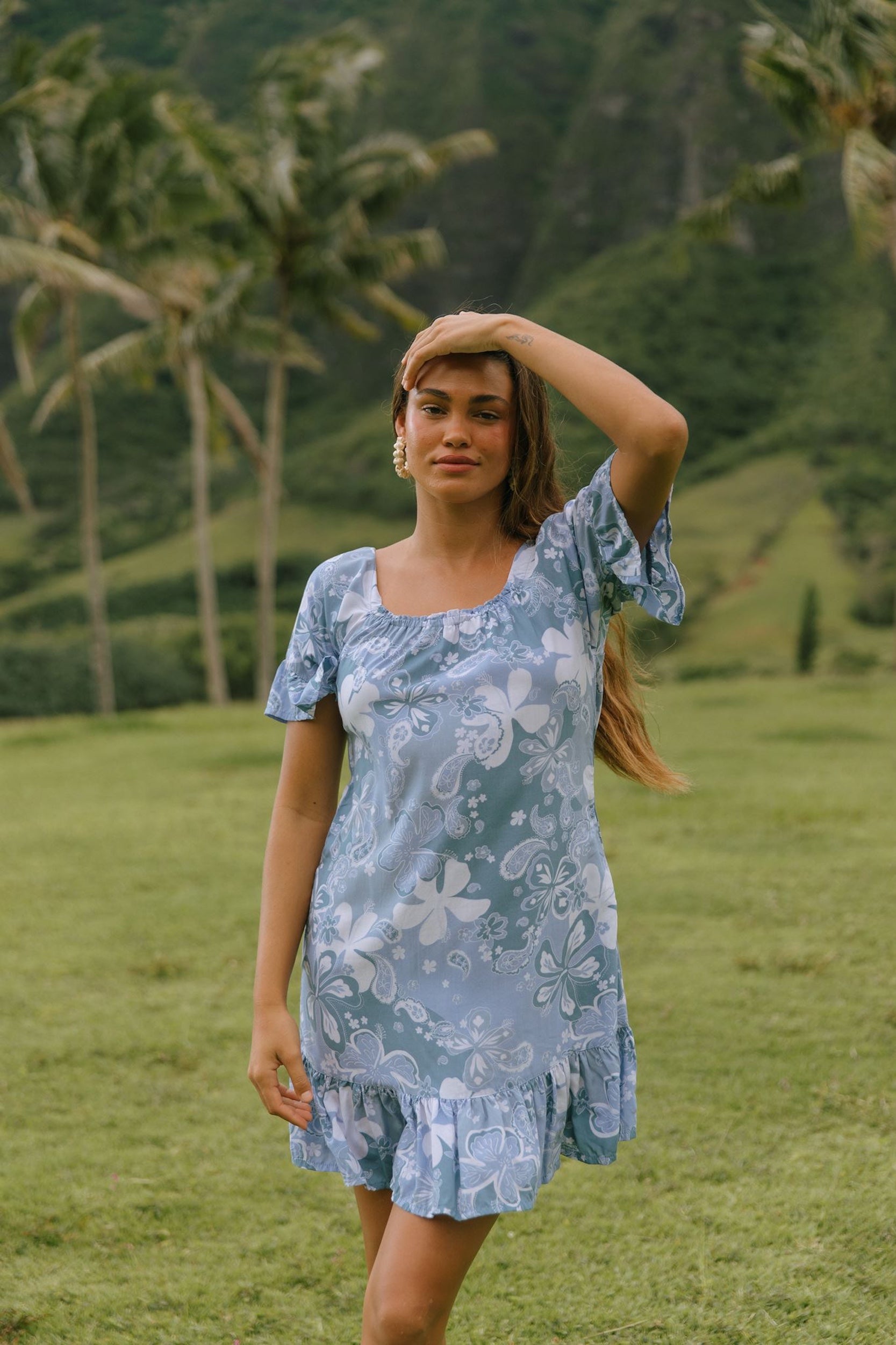 woman with brown hair wearing a blue paniolo printed mu'u style dress with flutter sleeves and a ruffle hem 