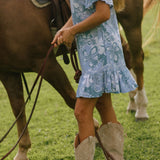 woman with brown hair wearing a blue paniolo printed mu'u style dress with flutter sleeves and a ruffle hem 