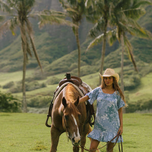 woman with brown hair wearing a blue paniolo printed mu'u style dress with flutter sleeves and a ruffle hem 