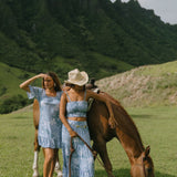 woman with brown hair wearing a blue paniolo printed mu'u style dress with flutter sleeves and a ruffle hem 