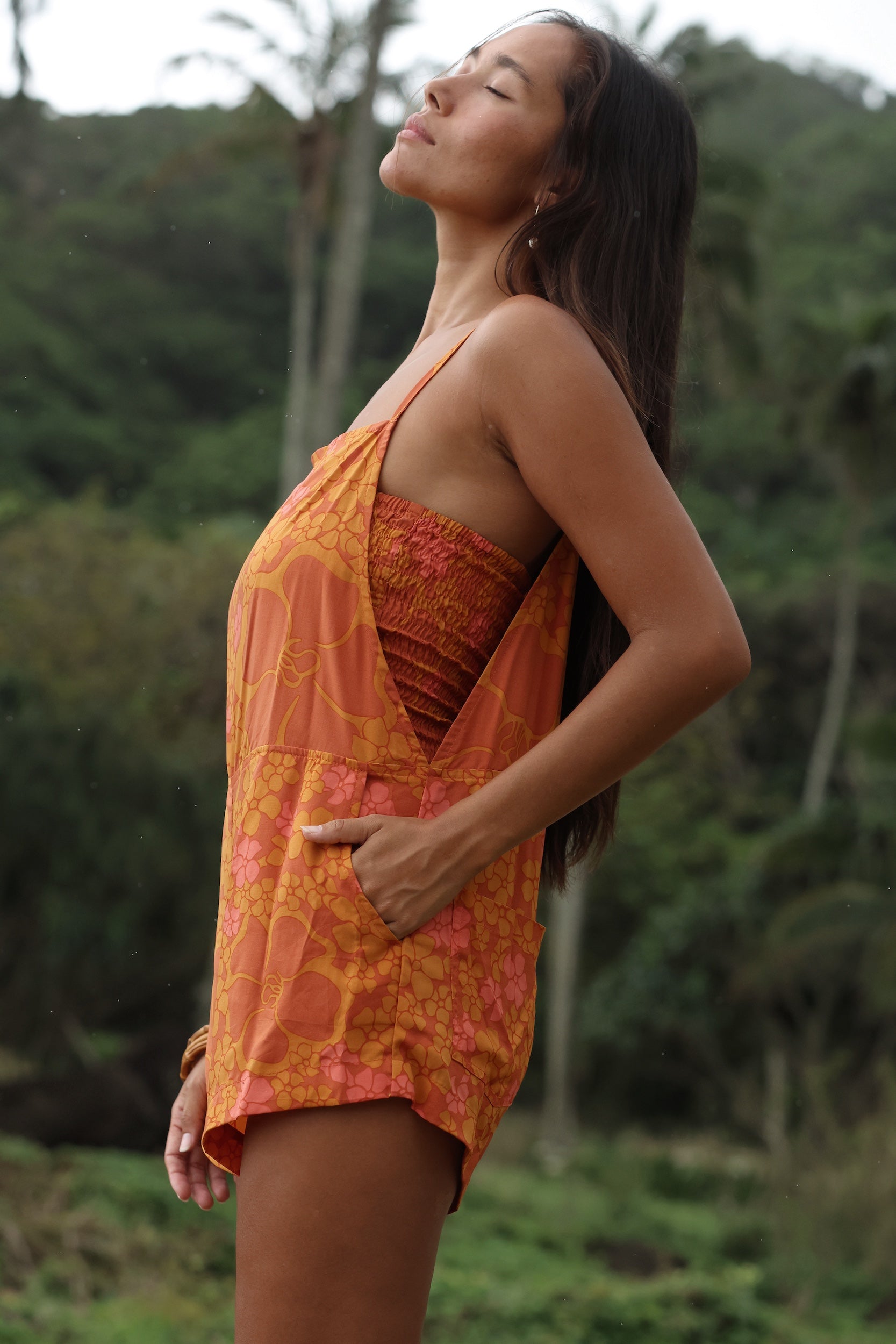 woman with brown hair wearing an orange overall style romper with rumi bandeau underneath with puakenikeni print