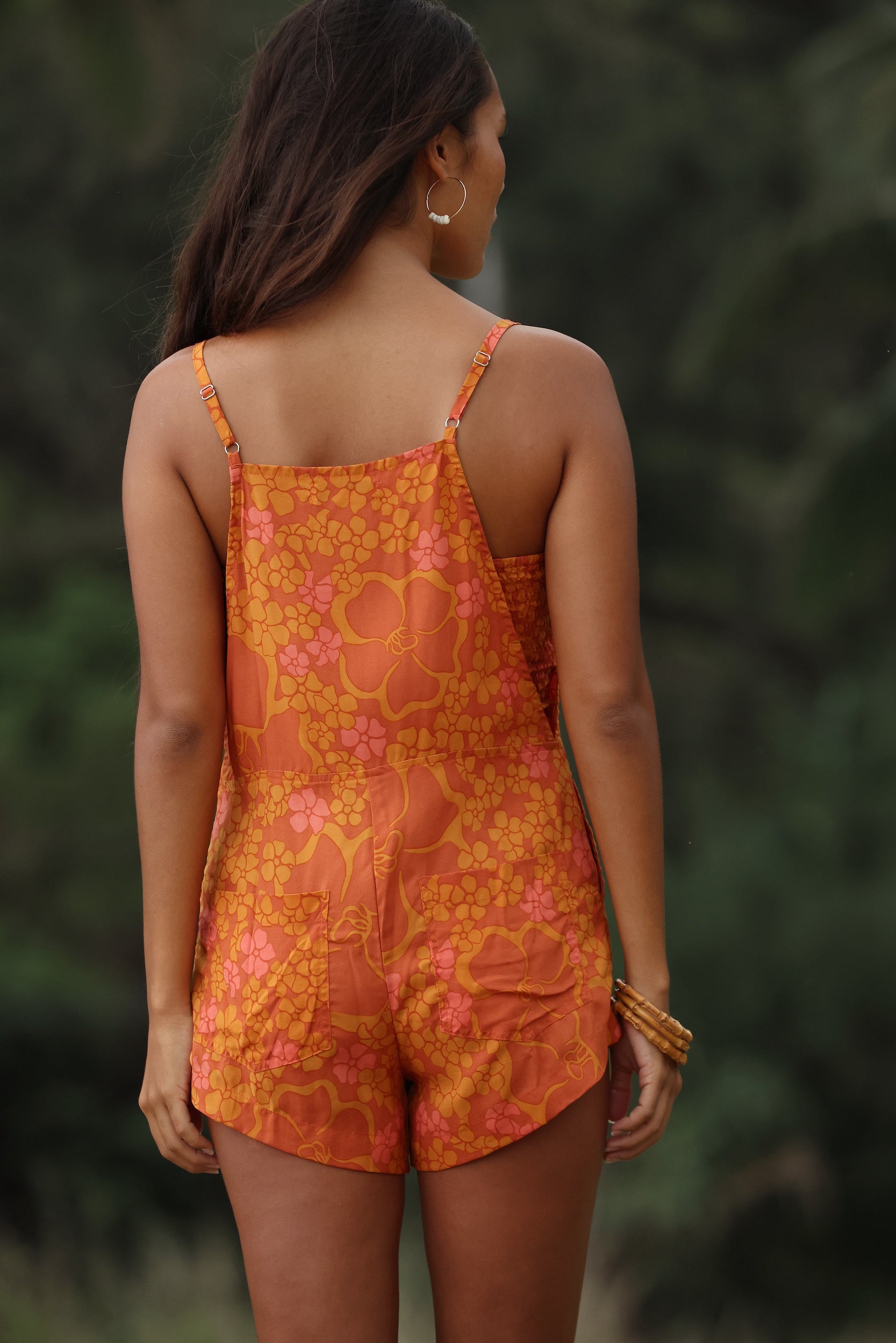 woman with brown hair wearing an orange overall style romper with rumi bandeau underneath with puakenikeni print