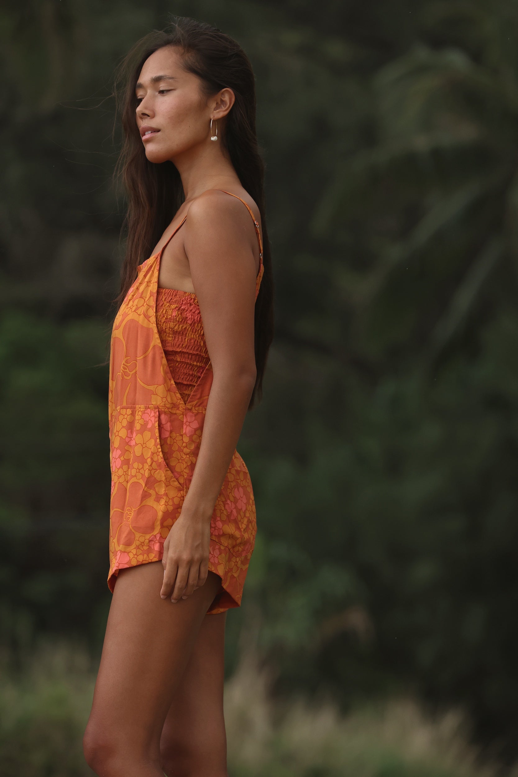 woman with brown hair wearing an orange overall style romper with rumi bandeau underneath with puakenikeni print