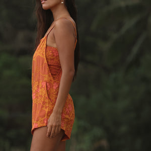 woman with brown hair wearing an orange overall style romper with rumi bandeau underneath with puakenikeni print