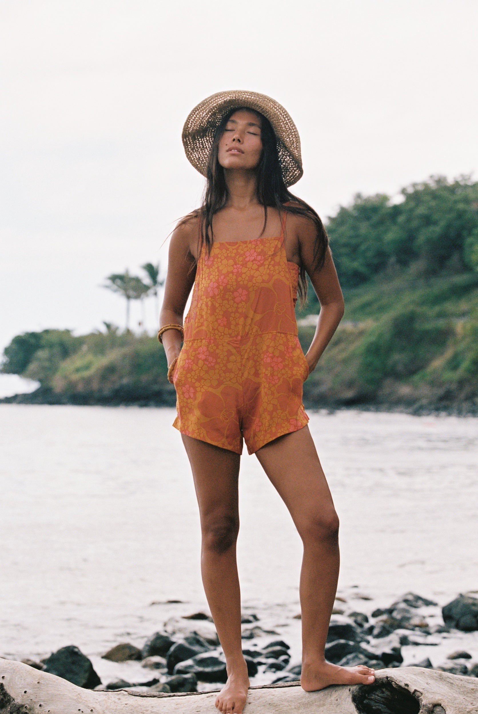 woman with brown hair wearing an orange overall style romper with rumi bandeau underneath with puakenikeni print