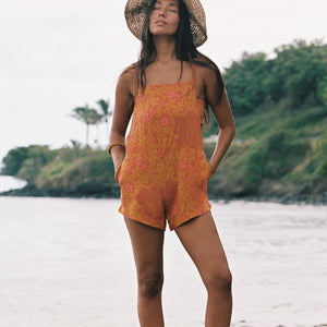 woman with brown hair wearing an orange overall style romper with rumi bandeau underneath with puakenikeni print
