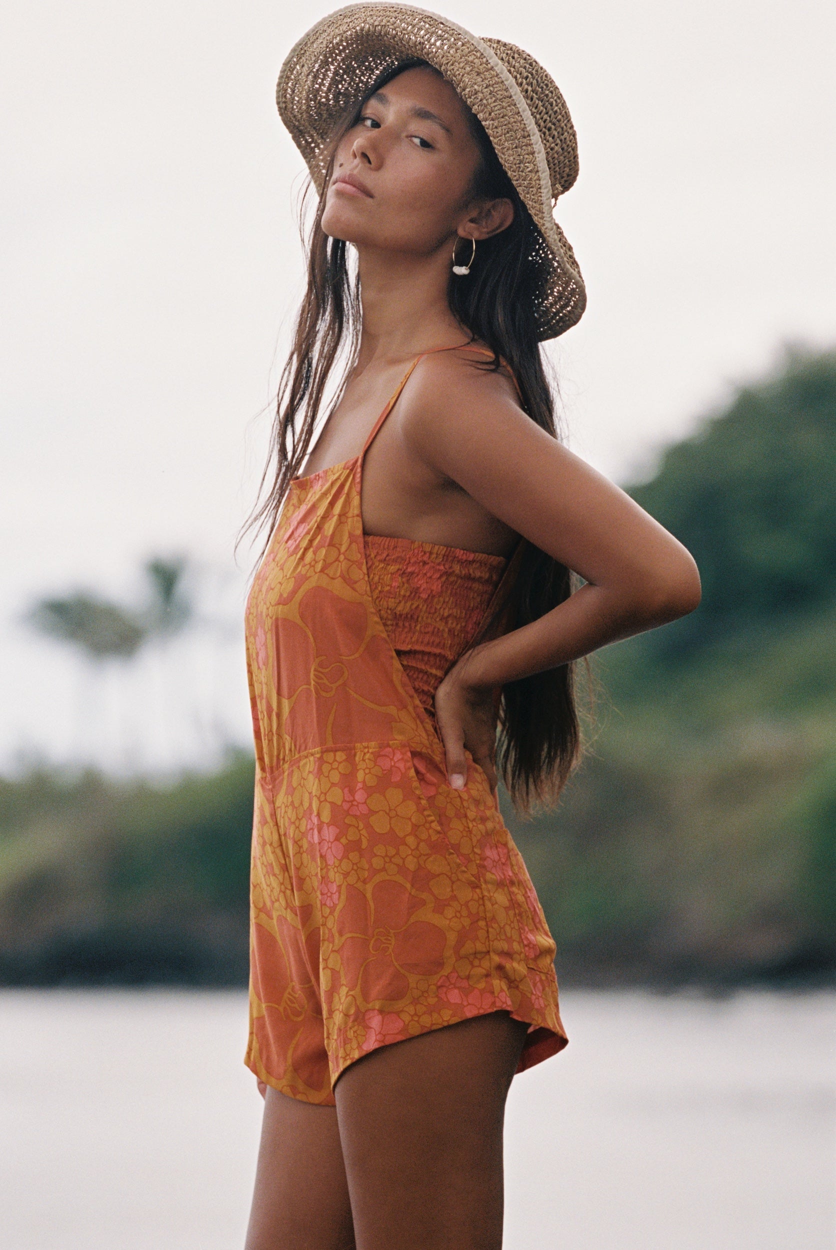 woman with brown hair wearing an orange overall style romper with rumi bandeau underneath with puakenikeni print