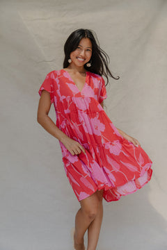 woman with brown hair wearing a red and pink v neck flowy knee length dress with mao flower pattern
