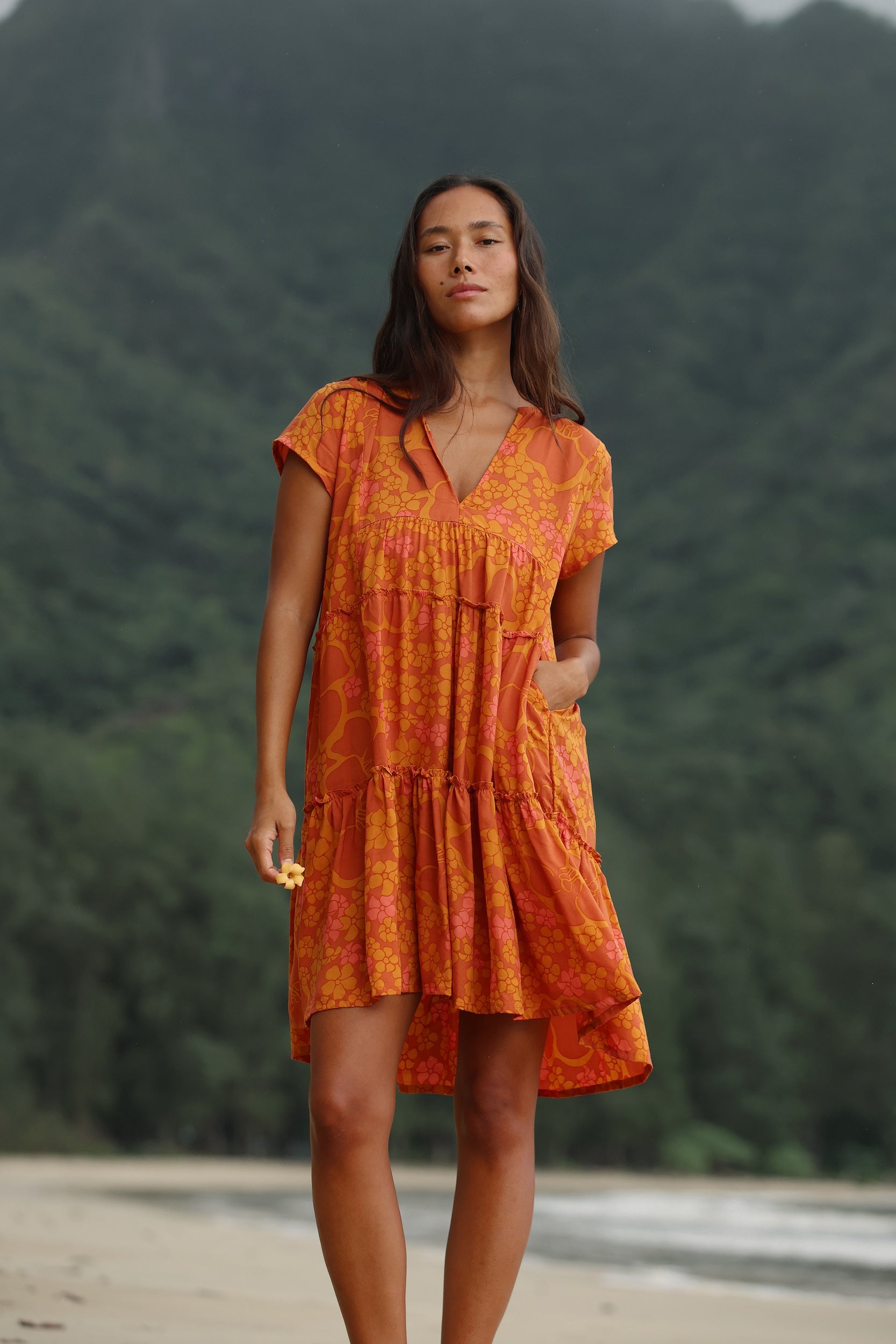 woman with brown hair wearing an orange v neck flowy over sized dress with puakenikeni print