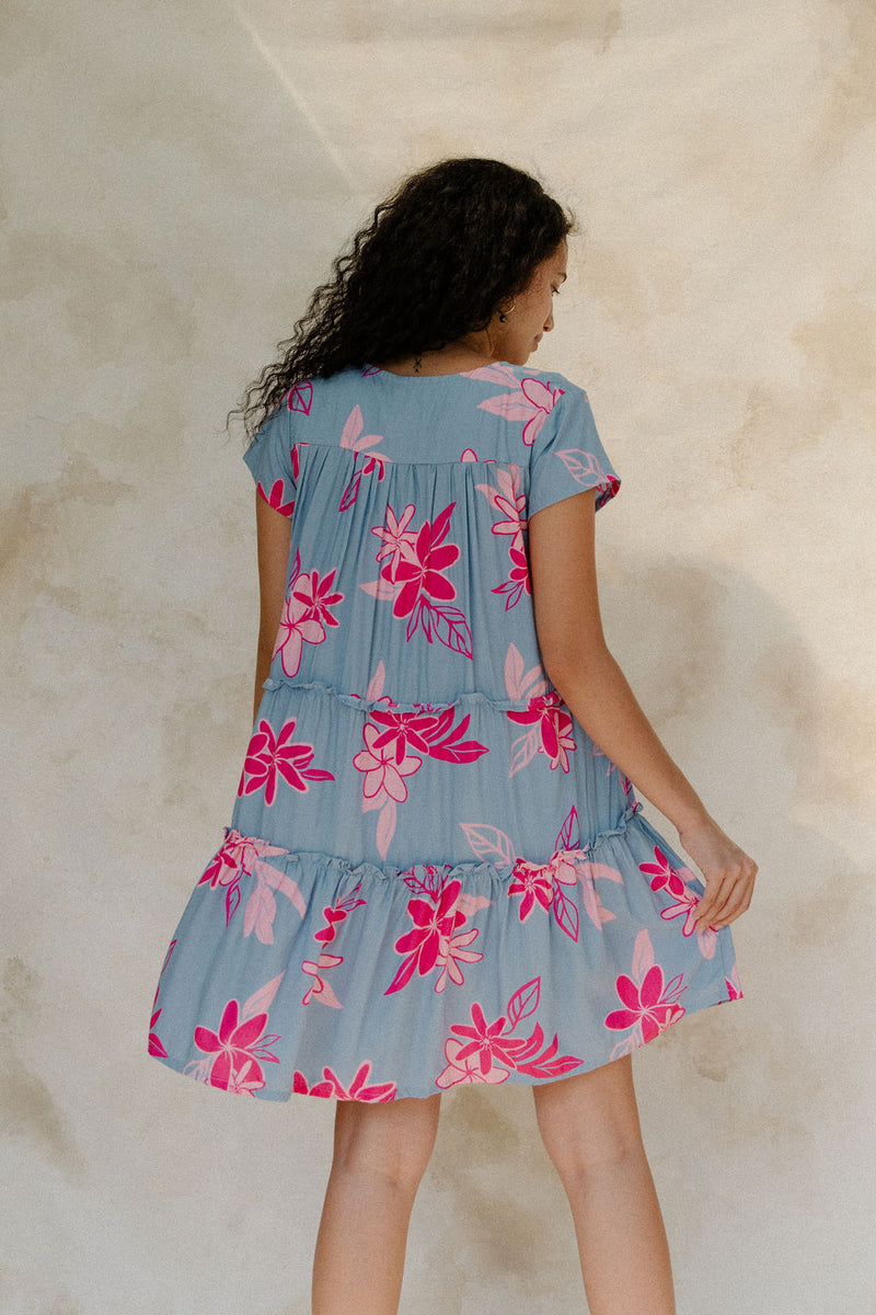 Woman in short sleeve flowy tiered dress with pink floral on light blue ground.