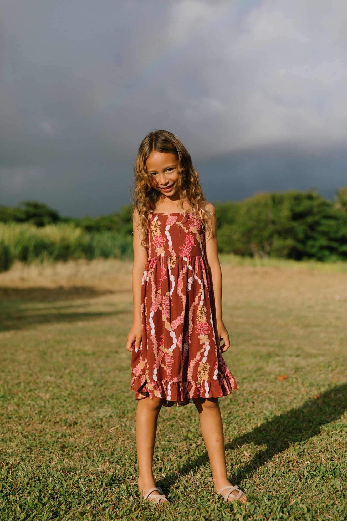 little girl wearing a red-brown tie back dress with ruffle hem and lei print