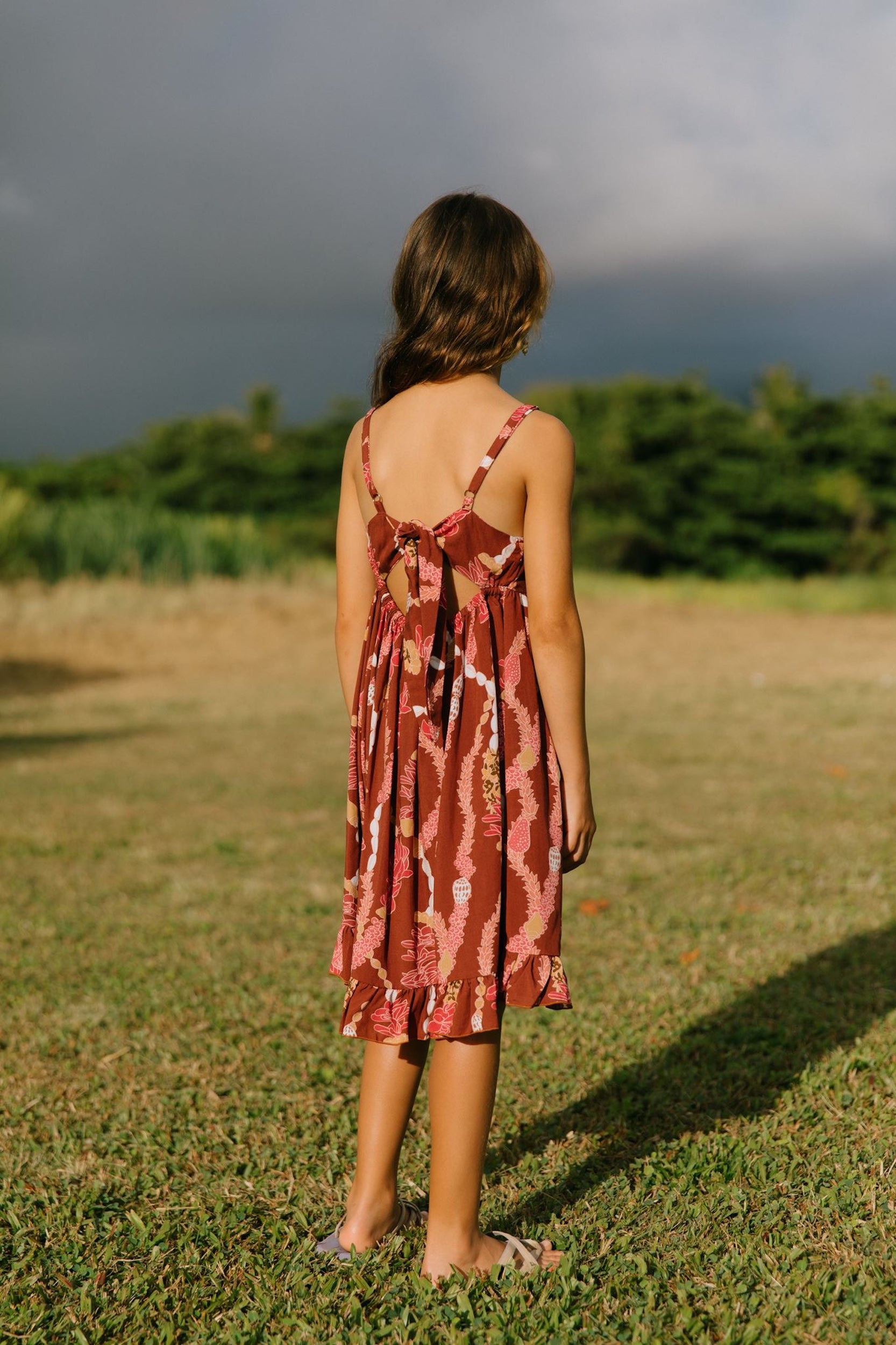 little girl wearing a red-brown tie back dress with ruffle hem and lei print