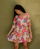 Woman wearing flowy tiered dress with 3/4 sleeves and multi colored floral print on light pink ground.