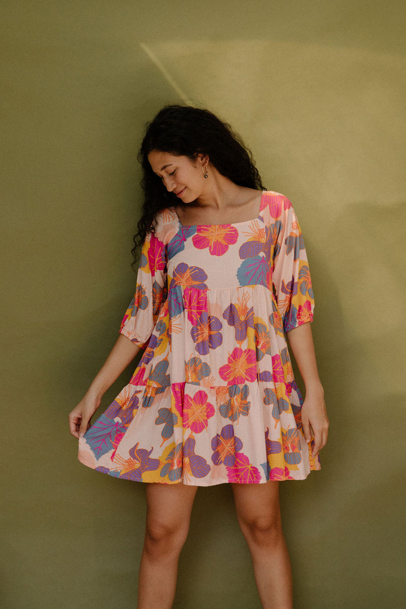 Woman wearing flowy tiered dress with 3/4 sleeves and multi colored floral print on light pink ground.