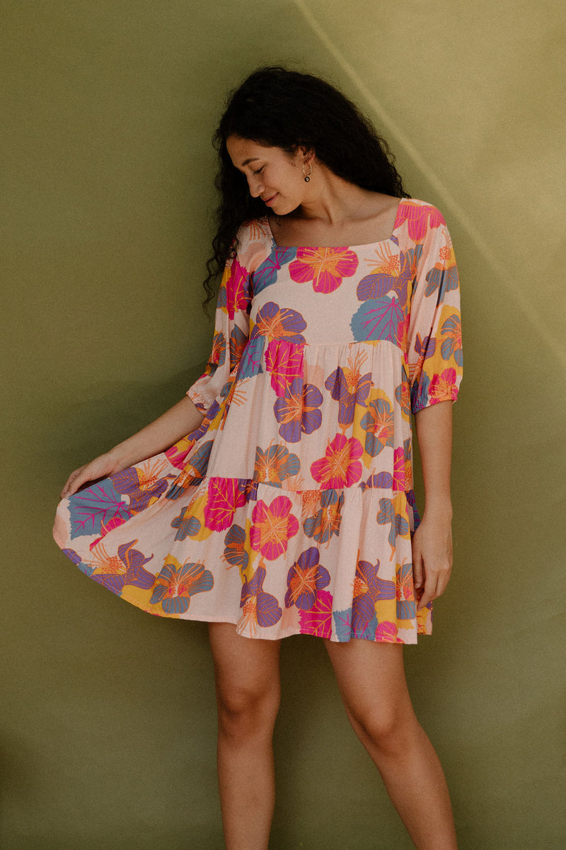 Woman wearing flowy tiered dress with 3/4 sleeves and multi colored floral print on light pink ground.