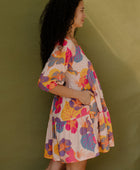 Woman wearing flowy tiered dress with 3/4 sleeves and multi colored floral print on light pink ground.