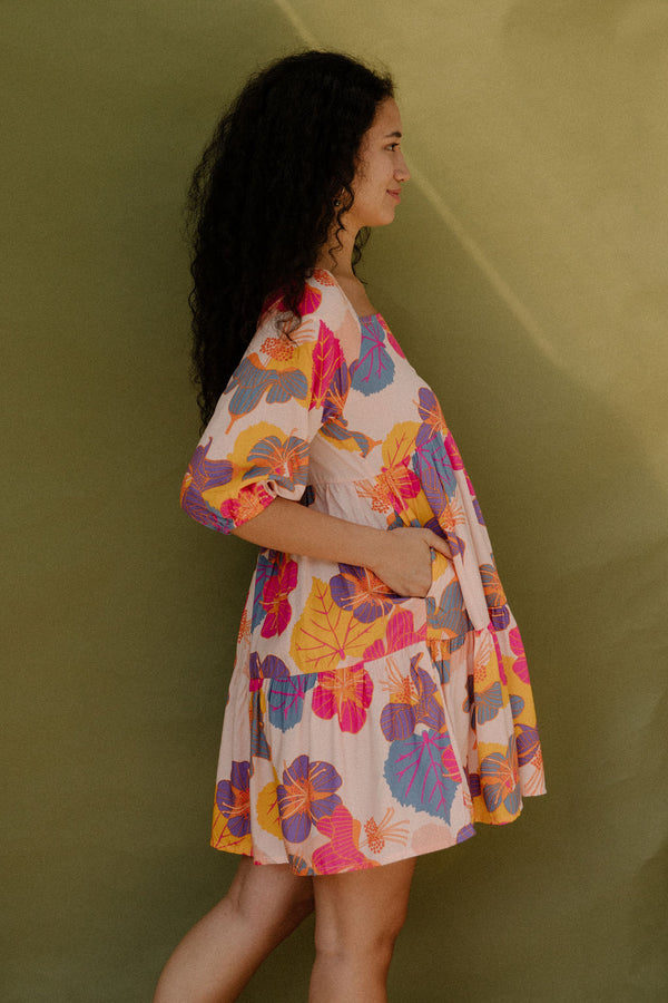 Woman wearing flowy tiered dress with 3/4 sleeves and multi colored floral print on light pink ground.