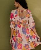 Woman wearing flowy tiered dress with 3/4 sleeves and multi colored floral print on light pink ground.