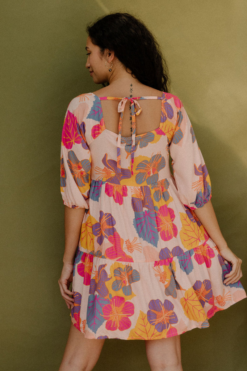 Woman wearing flowy tiered dress with 3/4 sleeves and multi colored floral print on light pink ground.