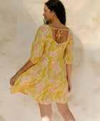 Woman in short flowy tiered dress with 3/4 sleeves in white and peach floral print with yellow ground.