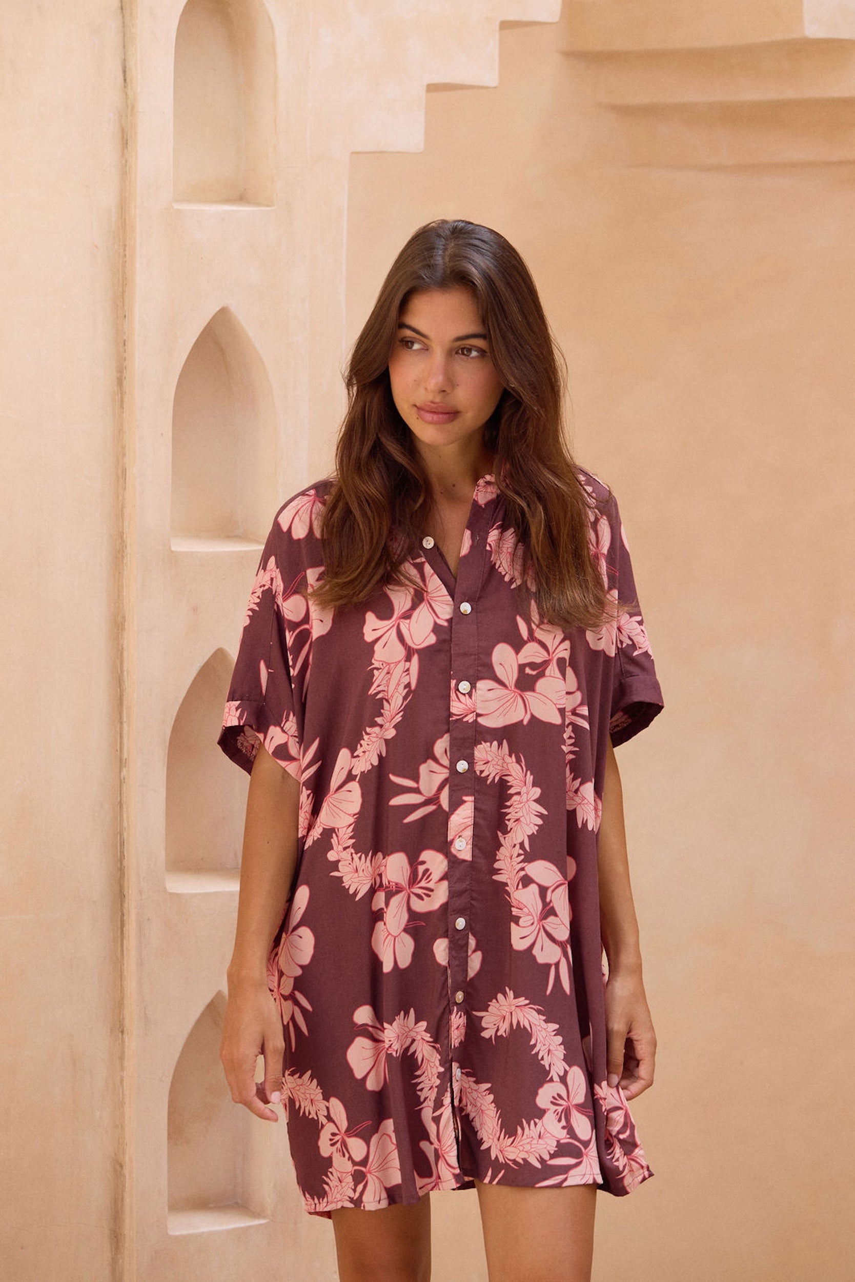 woman with brown hair wearing.a brown/maroon button up dress with pink lei awapuhi print