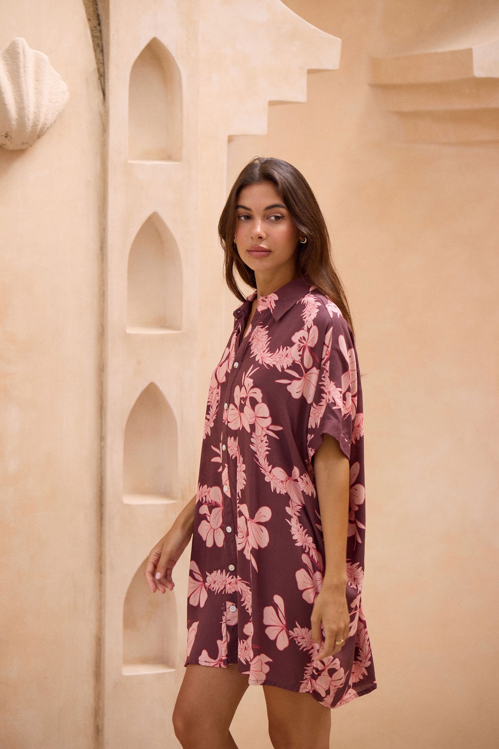 woman with brown hair wearing.a brown/maroon button up dress with pink lei awapuhi print