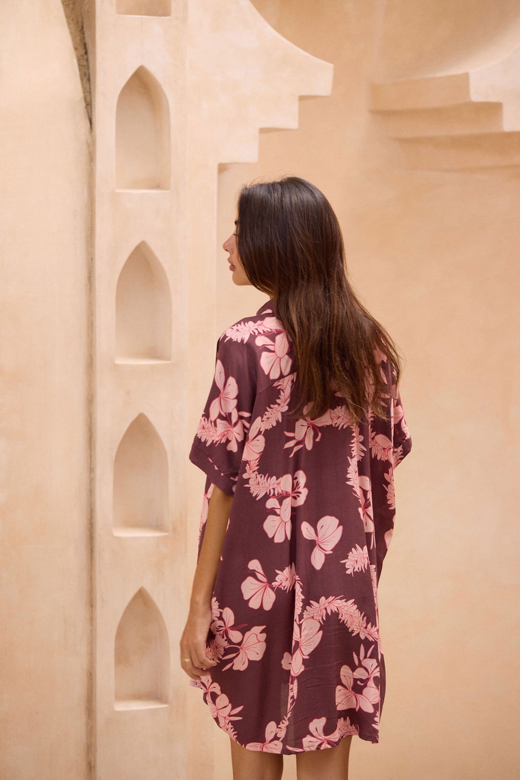 woman with brown hair wearing.a brown/maroon button up dress with pink lei awapuhi print