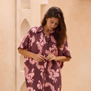 woman with brown hair wearing.a brown/maroon button up dress with pink lei awapuhi print