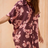 woman with brown hair wearing.a brown/maroon button up dress with pink lei awapuhi print