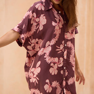 woman with brown hair wearing.a brown/maroon button up dress with pink lei awapuhi print