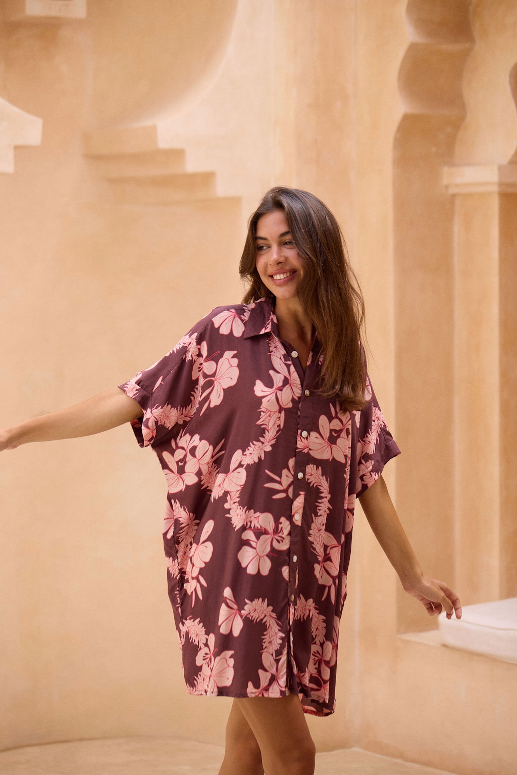woman with brown hair wearing.a brown/maroon button up dress with pink lei awapuhi print