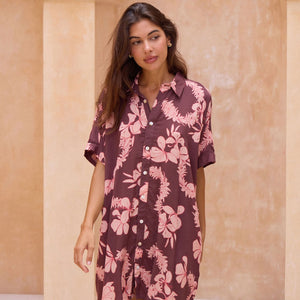 woman with brown hair wearing.a brown/maroon button up dress with pink lei awapuhi print