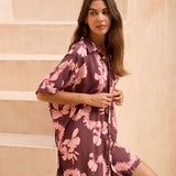 woman with brown hair wearing.a brown/maroon button up dress with pink lei awapuhi print