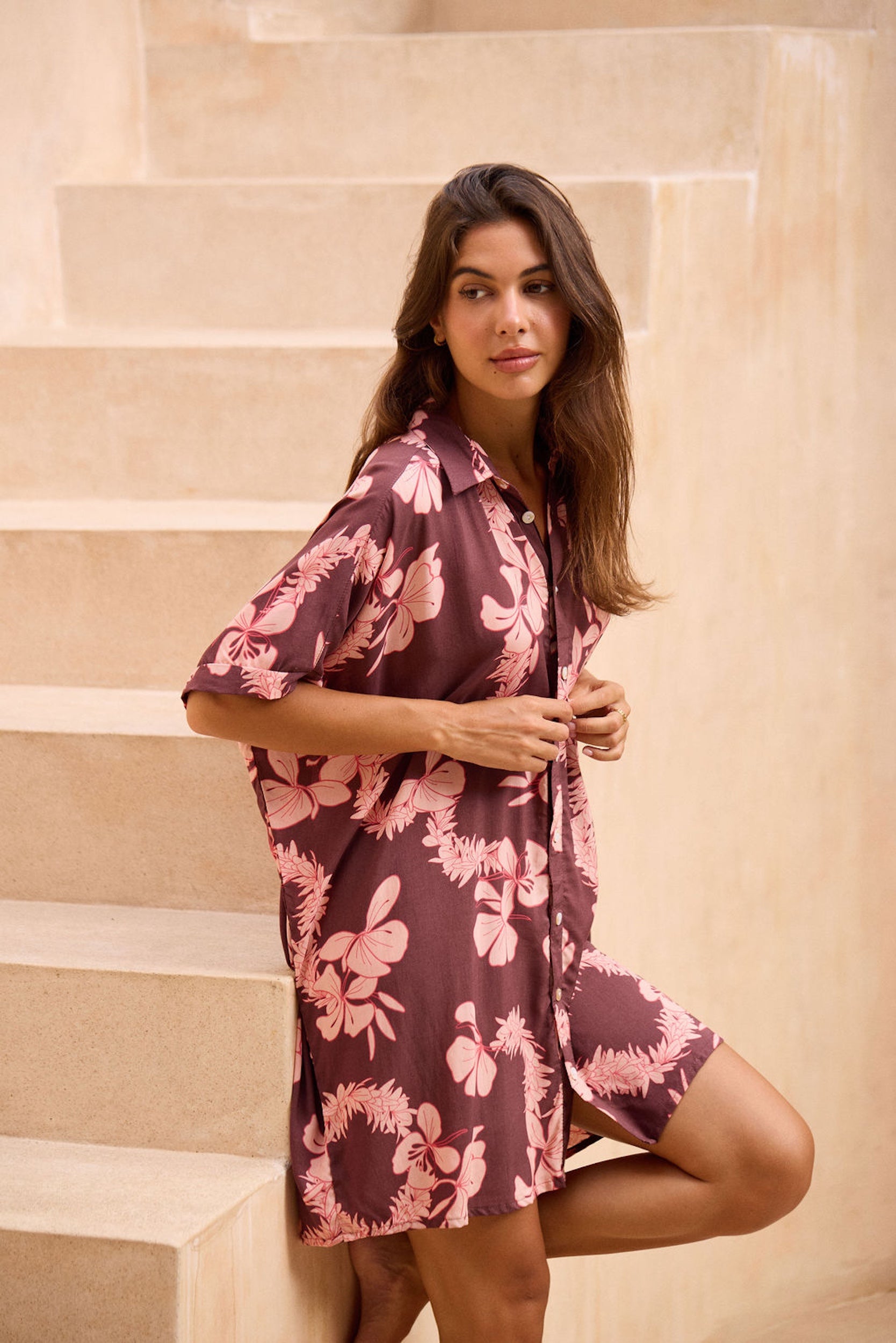woman with brown hair wearing.a brown/maroon button up dress with pink lei awapuhi print